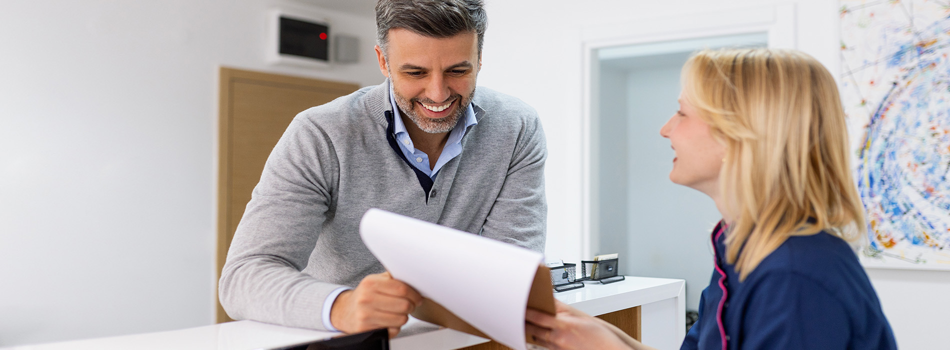 The image shows two individuals, likely professionals, engaged in a conversation at a desk with papers, set against a blurred office background.