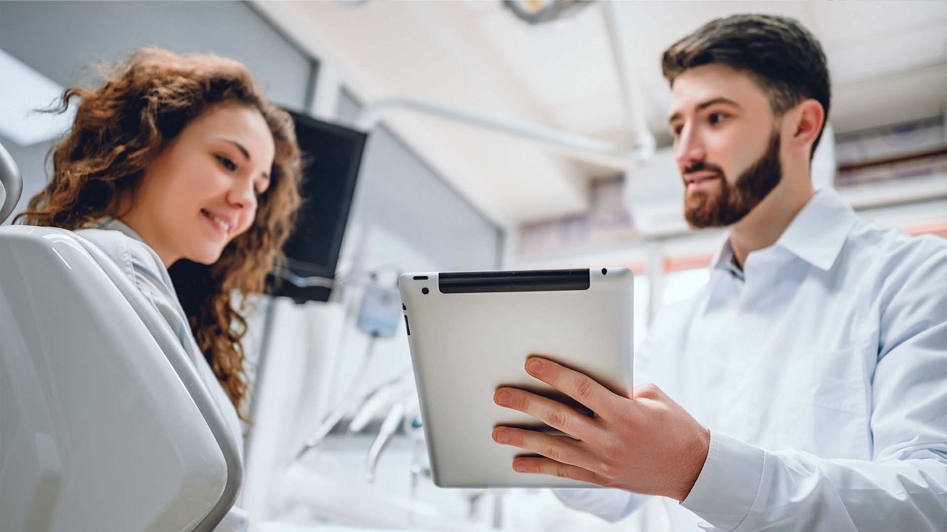 A man and woman in a medical setting, with the man holding a tablet, looking at something on it while the woman stands beside him.