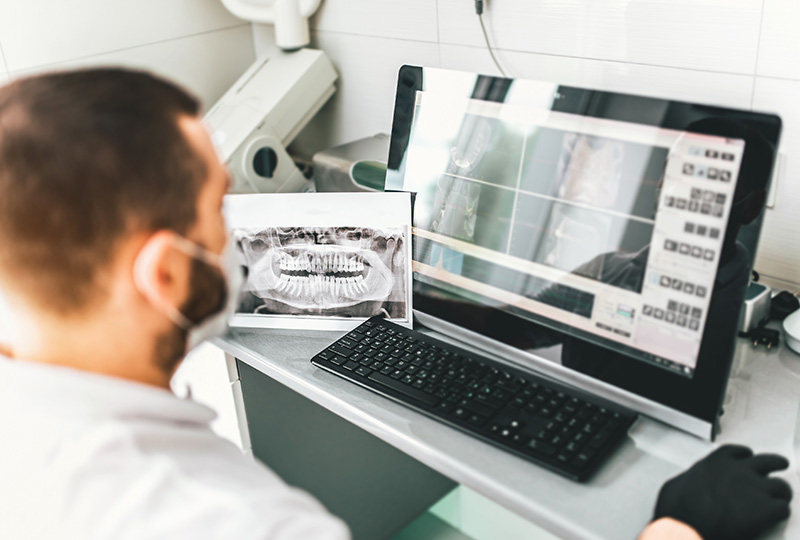 The image shows a person sitting at a computer monitor with medical equipment behind them, displaying an X-ray image on the screen.
