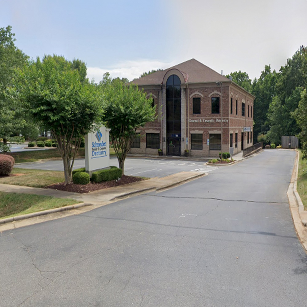 The image shows a two-story building with a brick facade, a sign indicating SCHOOL OF NURSING on its side, and a parking lot with trees and a paved driveway leading up to it.