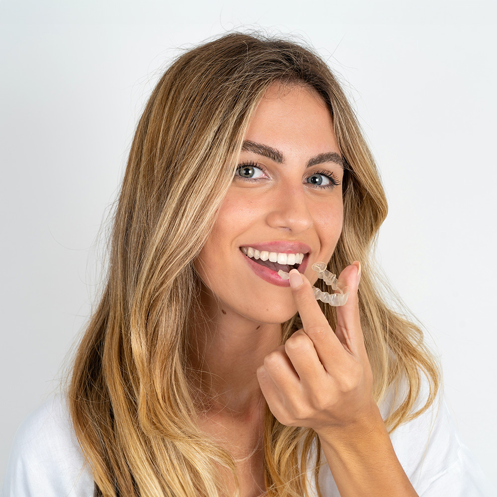 A woman with long blonde hair is holding a white toothbrush near her mouth.