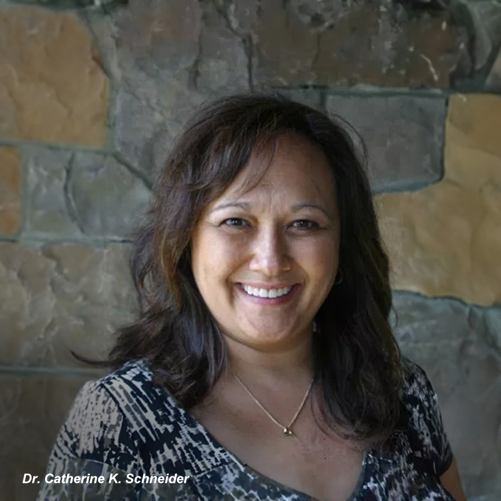 The image shows a smiling woman with long hair standing outdoors against a natural backdrop, posing for a portrait.