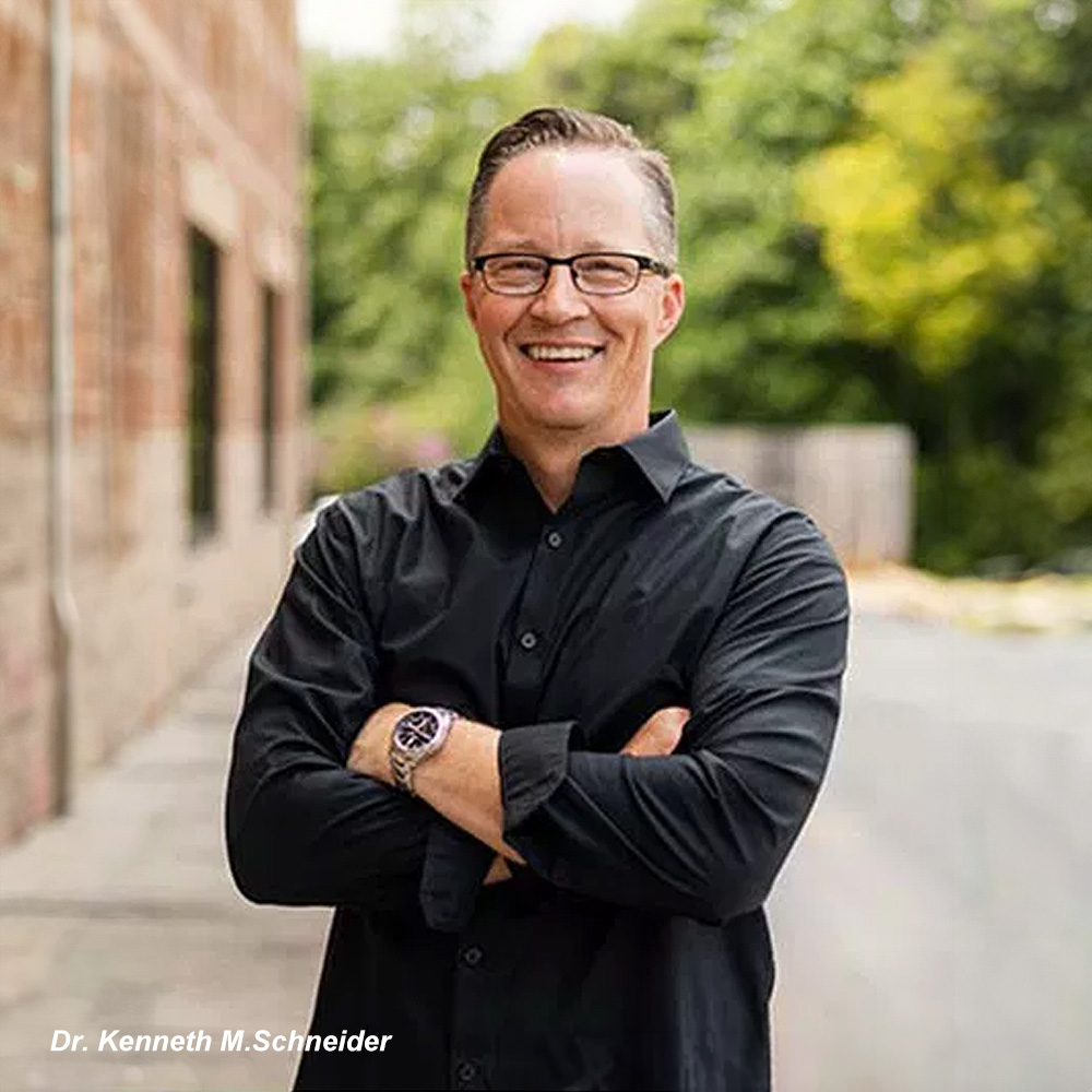 The image shows a man standing outdoors with his arms crossed, wearing glasses, a black shirt, and a watch, against a backdrop of a building with a brick wall and a tree. He appears to be posing for a portrait.