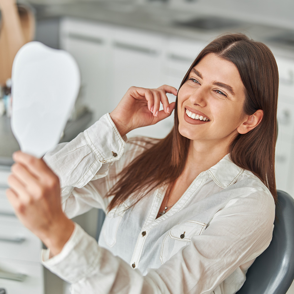 Woman seated at dental chair, smiling, holding up mirror towards her face while looking into it.